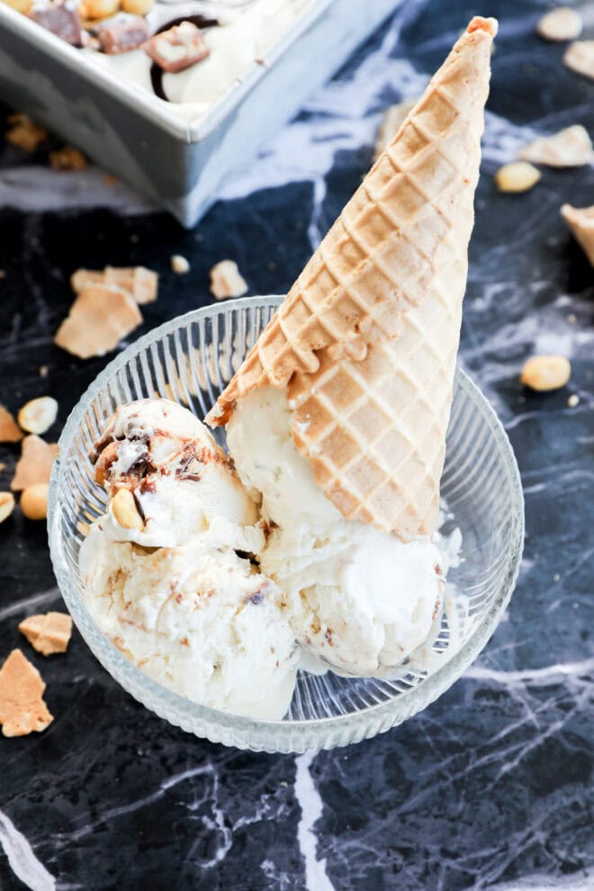 A glass bowl with scoops of Snickers Ice Cream and a waffle cone placed upside down on top, set on a black marble surface with pieces of nuts and cone scattered around.