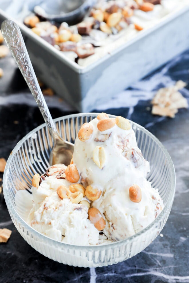 A glass bowl with two scoops of Snickers Ice Cream topped with peanuts, with a silver spoon inside. A loaf pan filled with more creamy ice cream and nuts sits in the background on a dark marble surface.