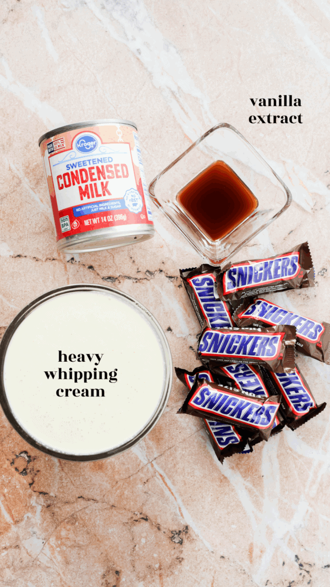 Top-down view of ingredients for Snickers Ice Cream on a marble surface: a can of sweetened condensed milk, a small glass dish of vanilla extract, a bowl of heavy whipping cream, and several mini Snickers bars.