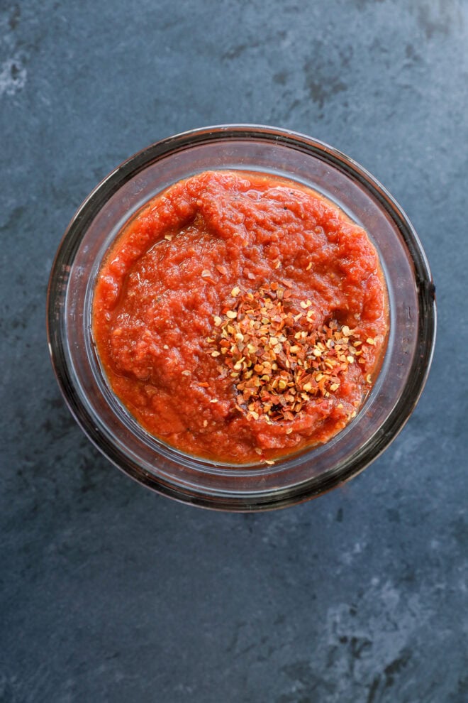 A glass bowl filled with thick red tomato sauce, perfect for spicy pizza, is topped with a sprinkle of crushed red pepper flakes and sits on a dark gray surface.