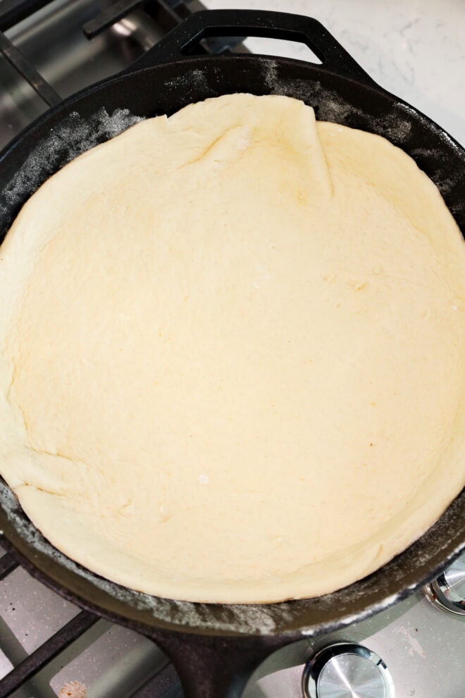 Unbaked pizza dough pressed evenly into a black cast iron skillet, resting on a stovetop with metal burner knobs visible.