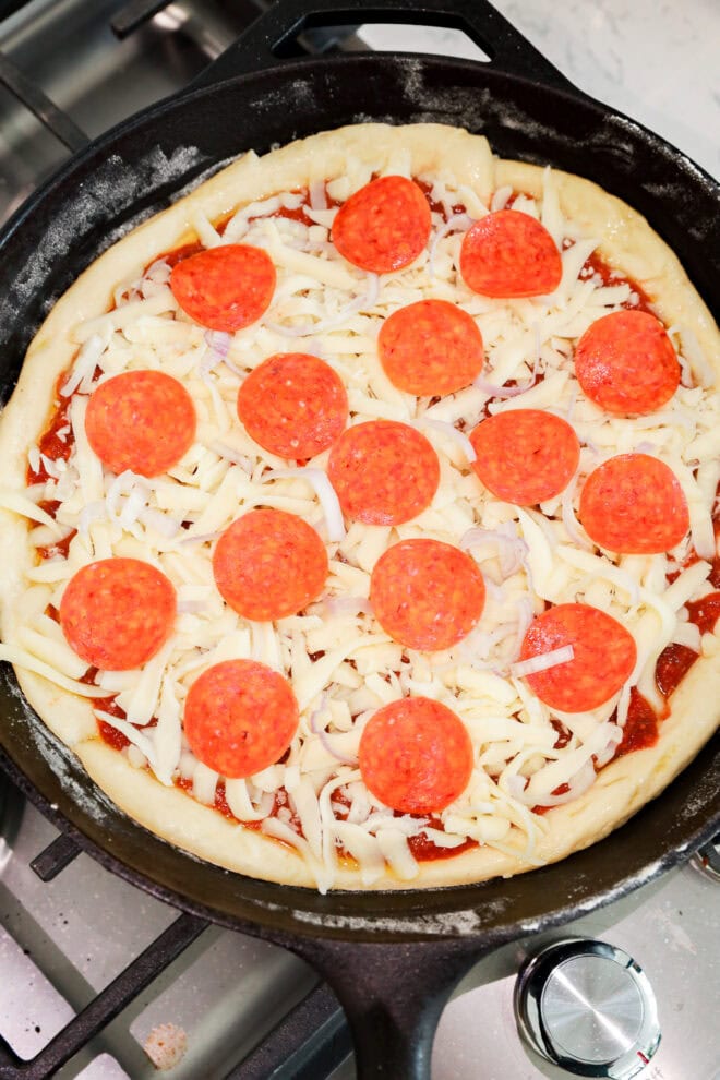 A cast iron skillet on a stove holds an uncooked spicy pizza, topped with shredded mozzarella cheese and pepperoni slices layered over zesty tomato sauce.