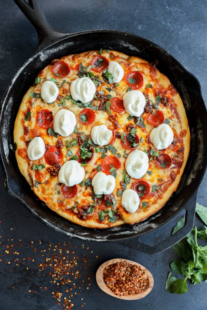 A freshly baked spicy pizza topped with pepperoni, melted cheese, basil, and dollops of ricotta in a black cast-iron skillet, with red pepper flakes and basil leaves on the dark surface nearby.