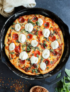 A freshly baked spicy pizza in a cast-iron skillet, topped with melted mozzarella cheese, pepperoni, and fresh basil, with a bowl of red pepper flakes and basil leaves nearby.