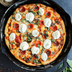 A freshly baked spicy pizza in a cast-iron skillet, topped with melted mozzarella cheese, pepperoni, and fresh basil, with a bowl of red pepper flakes and basil leaves nearby.