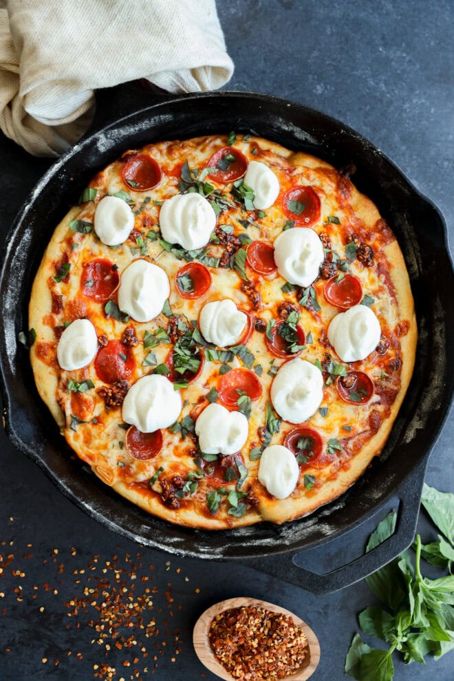 A freshly baked spicy pizza in a cast-iron skillet, topped with melted mozzarella cheese, pepperoni, and fresh basil, with a bowl of red pepper flakes and basil leaves nearby.