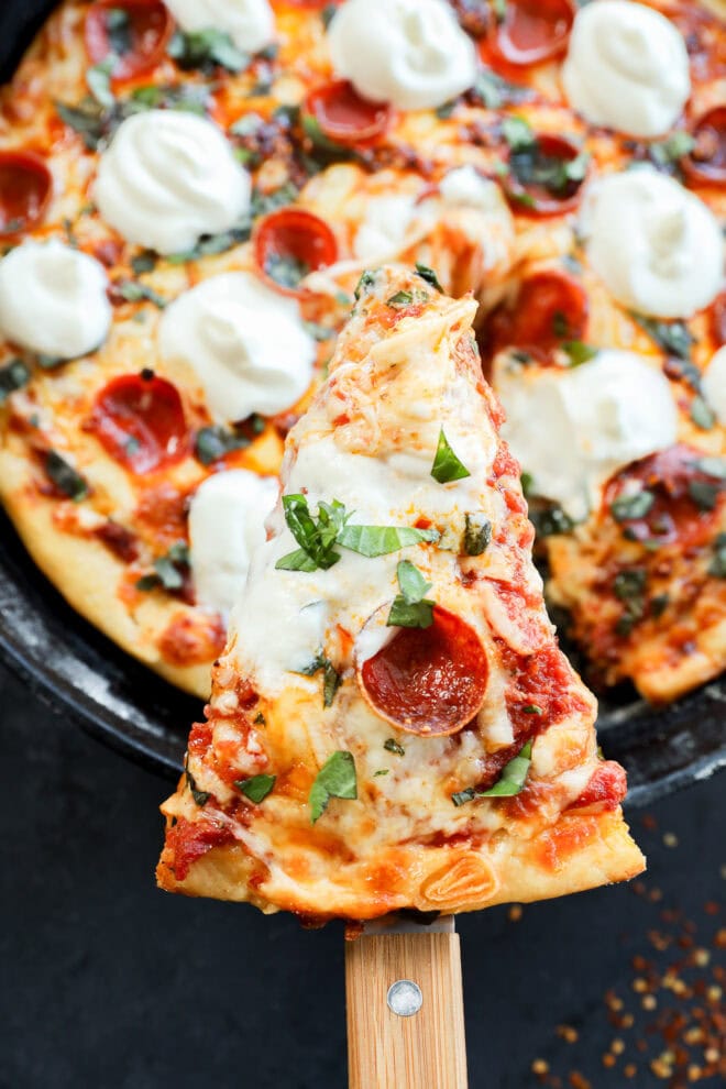 A close-up of a spicy pizza with pepperoni, melted mozzarella, fresh basil, and tomato sauce. A single slice is being lifted from the whole pizza with a wooden spatula.