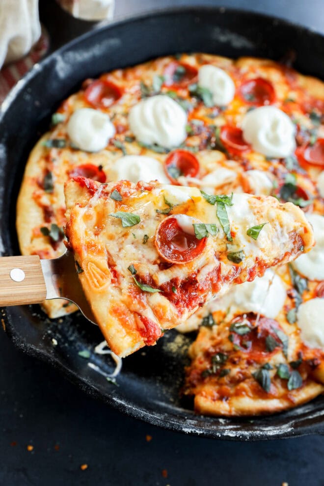 A close-up of a spicy pizza slice being lifted from a cast iron skillet, topped with melted cheese, fresh basil, and dollops of creamy white sauce. The rest of the pizza is visible in the skillet.