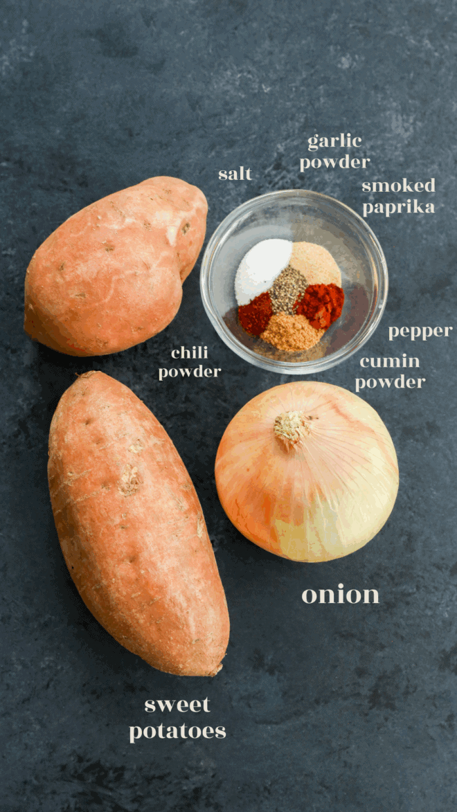 Two whole sweet potatoes, a peeled onion, and a small bowl containing salt, garlic powder, smoked paprika, chili powder, cumin powder, and pepper, all labeled, on a dark background.