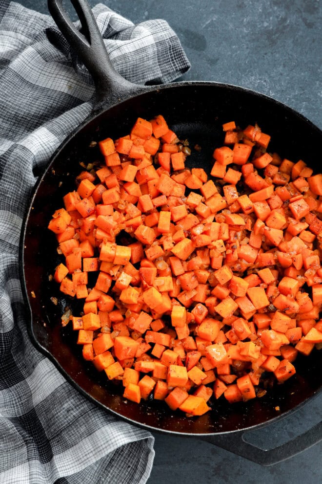 A cast iron skillet filled with diced, sautéed sweet potatoes sits on a dark surface next to a black and white plaid kitchen towel.