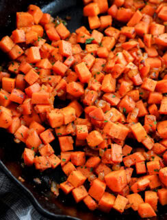Diced sweet potatoes sautéed with herbs and spices in a black skillet, showing a vibrant orange color and slightly crispy texture.