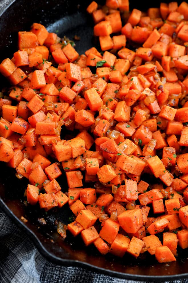 Diced sweet potatoes sautéed with herbs and spices in a black skillet, showing a vibrant orange color and slightly crispy texture.