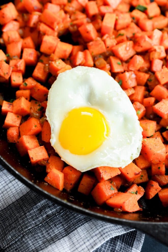 A fried egg with a bright yellow yolk sits on top of a skillet filled with diced, cooked sweet potatoes. The dish is garnished with herbs and rests on a striped cloth.