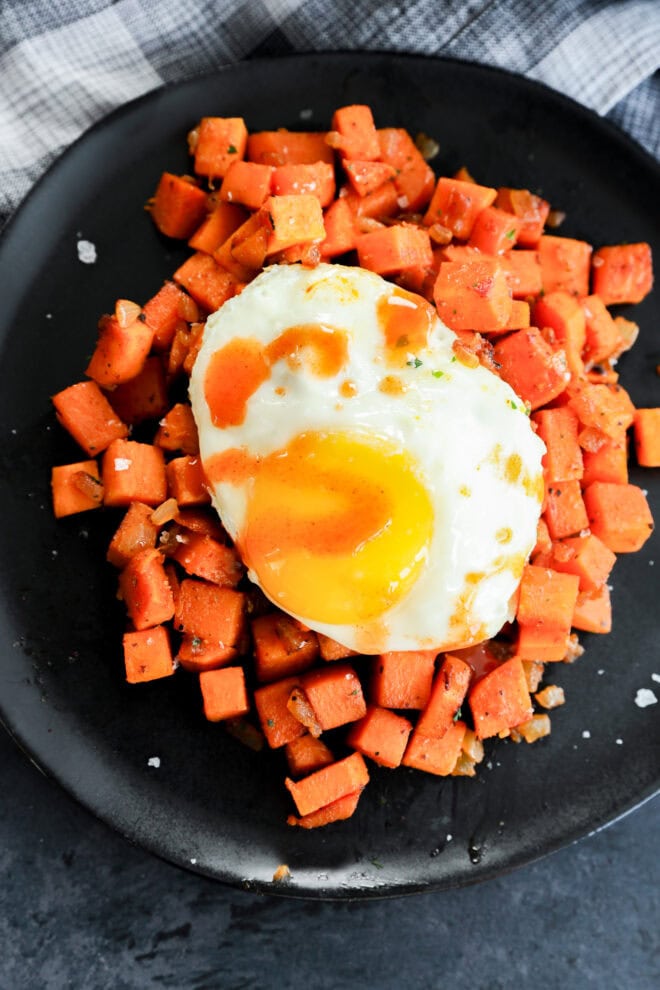 A black plate with a serving of diced, cooked sweet potatoes topped with a sunny-side-up egg and a drizzle of hot sauce. A black and white checkered cloth is visible in the background.