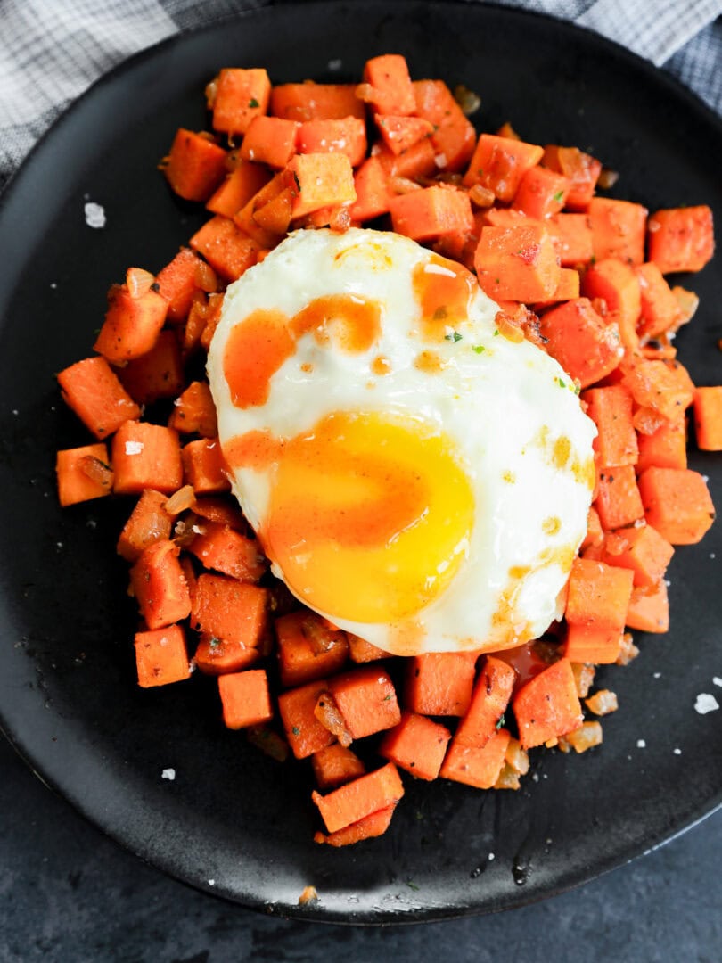A black plate with a serving of diced, cooked sweet potatoes topped with a sunny-side-up egg and a drizzle of hot sauce. A black and white checkered cloth is visible in the background.