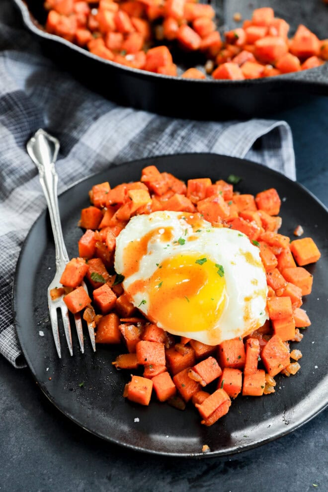 A black plate with diced sweet potatoes topped with a sunny-side-up egg and herbs, next to a fork on a plaid cloth, with a skillet of more sweet potatoes in the background.