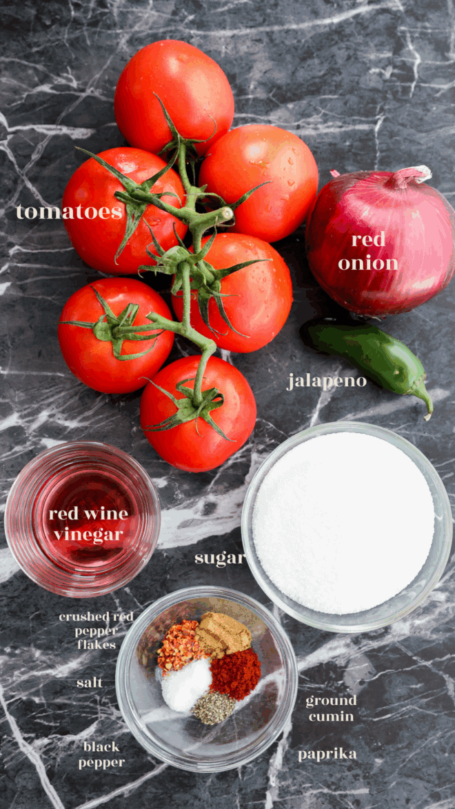 Fresh tomatoes on the vine, a red onion, a jalapeño, red wine vinegar, sugar, and various spices—all the essential ingredients for making vibrant Tomato Relish—are labeled and arranged on a dark marble surface.
