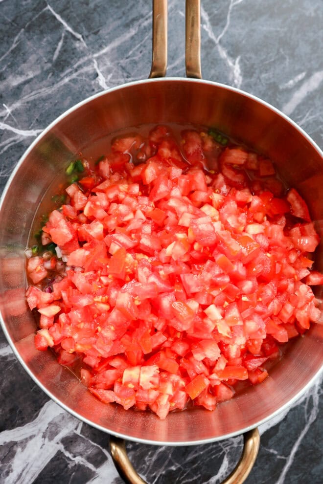 A copper pot filled with freshly diced tomatoes sits on a dark marble countertop.