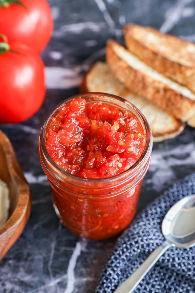 A glass jar filled with chunky tomato jam or tomato relish sits on a dark marble surface, surrounded by fresh tomatoes, slices of bread, a spoon, and a textured blue cloth.