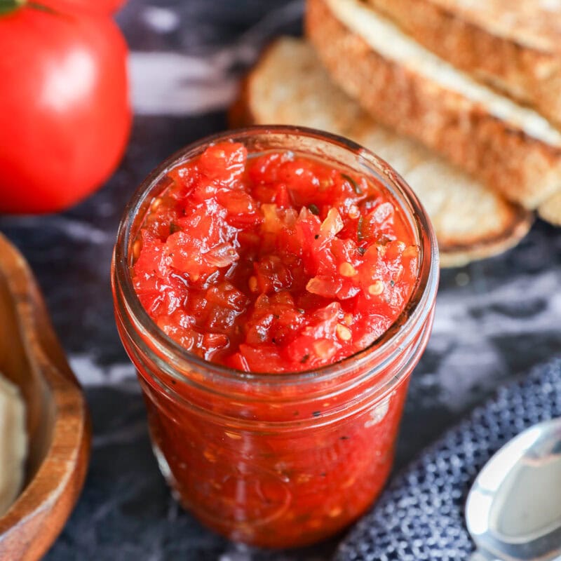 A glass jar filled with chunky tomato jam or tomato relish sits on a dark marble surface, surrounded by fresh tomatoes, slices of bread, a spoon, and a textured blue cloth.