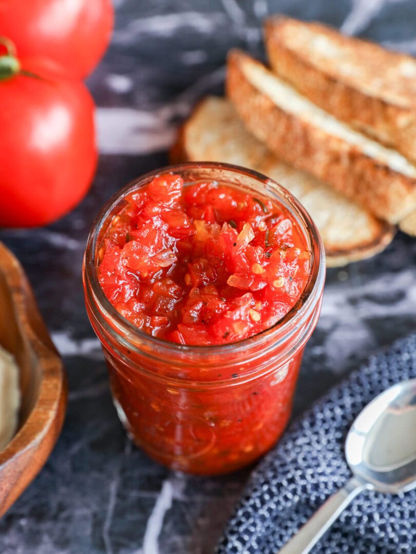 A glass jar filled with chunky tomato jam or tomato relish sits on a dark marble surface, surrounded by fresh tomatoes, slices of bread, a spoon, and a textured blue cloth.