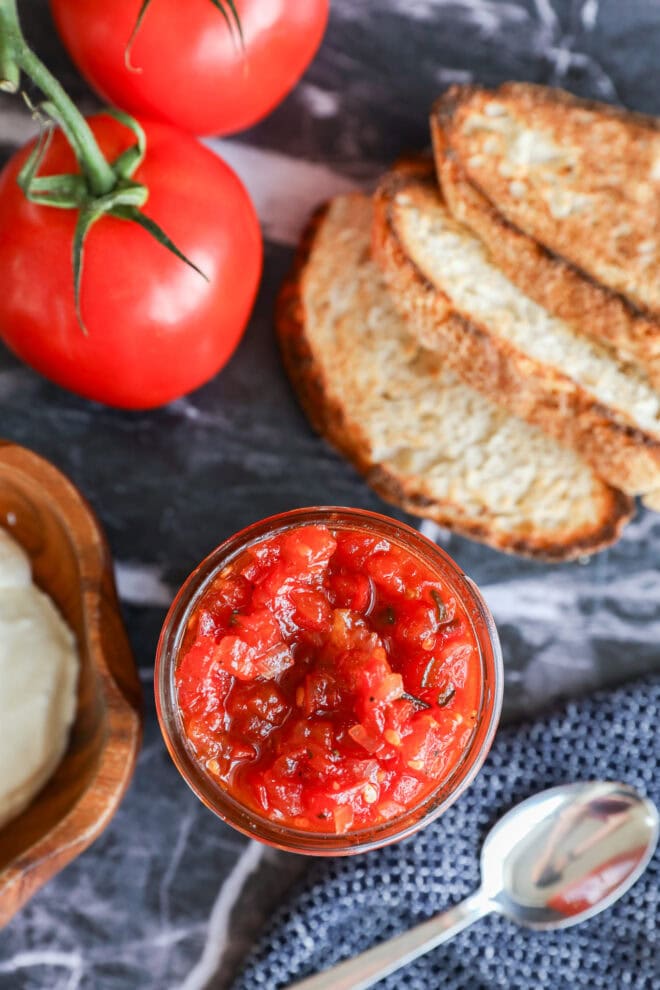 A jar of tomato relish sits on a marble surface next to slices of toasted bread, two fresh tomatoes on the vine, a bowl of white cheese, and a silver spoon.