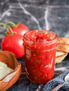 A glass jar filled with chunky tomato relish sits on a marble surface, surrounded by fresh tomatoes, slices of toasted bread, and a wooden bowl of butter.
