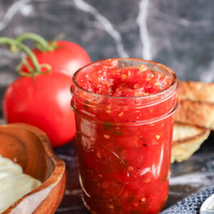 A glass jar filled with chunky tomato relish sits on a marble surface, surrounded by fresh tomatoes, slices of toasted bread, and a wooden bowl of butter.