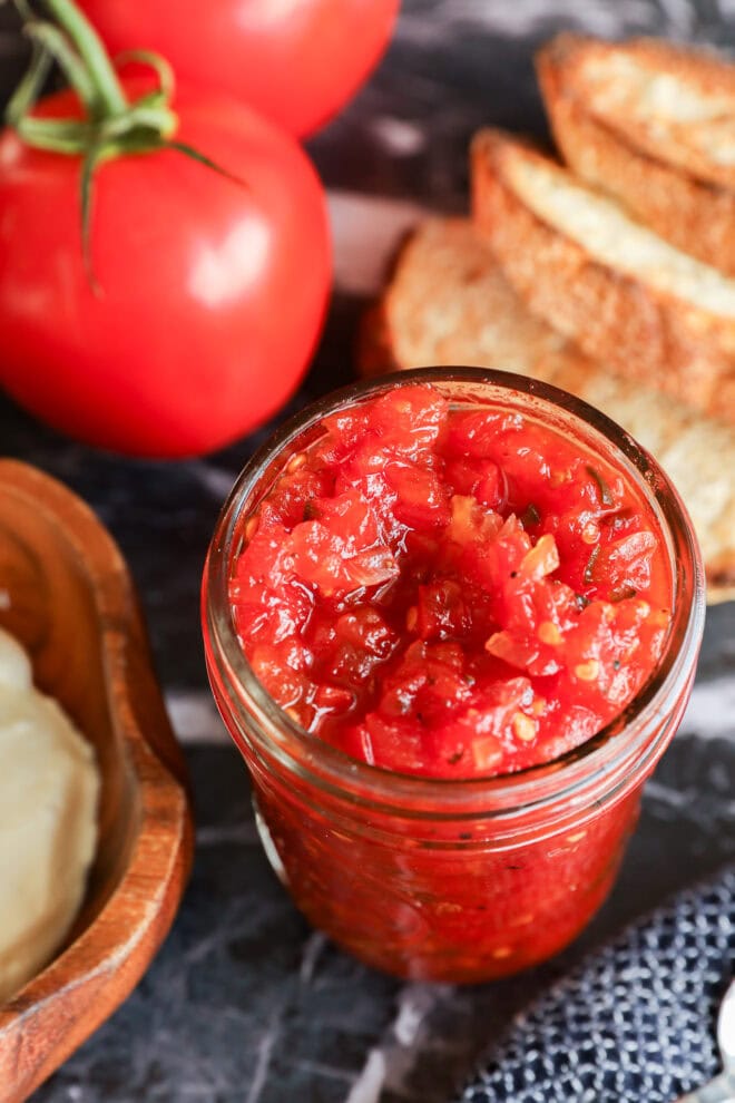 A glass jar filled with chunky tomato jam and tangy tomato relish sits on a table next to sliced bread, fresh tomatoes, and a wooden dish of butter.