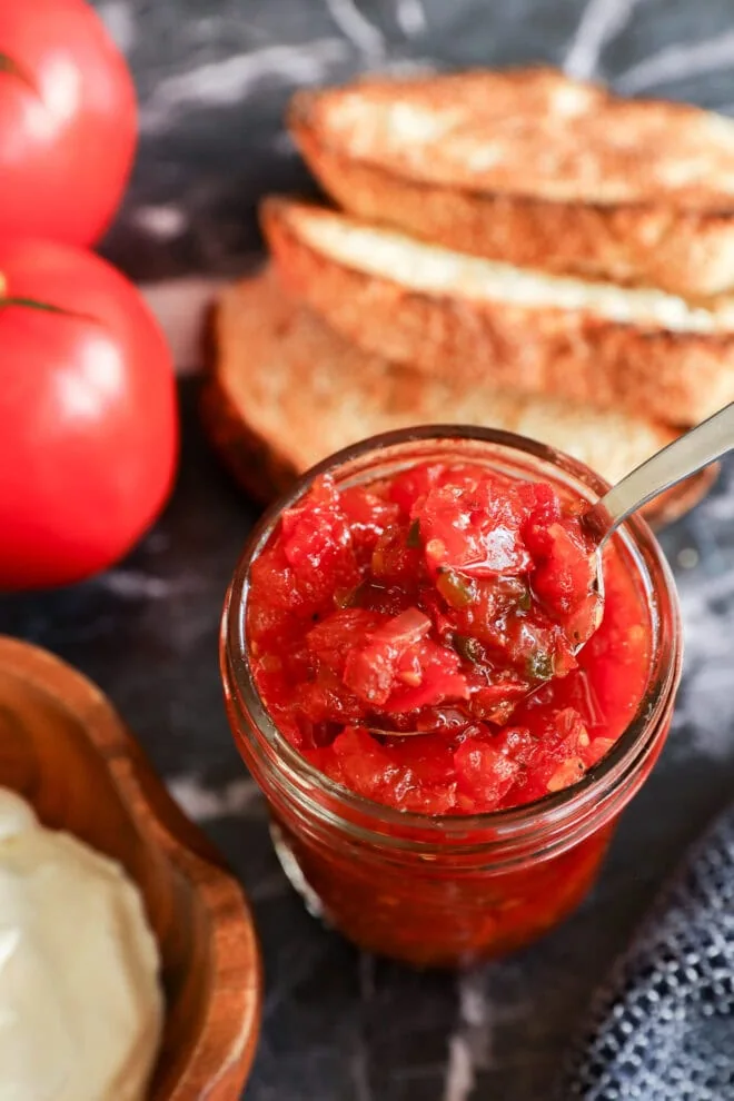 A jar of chunky tomato relish jam with a spoon inside, placed on a dark surface next to slices of toasted bread, fresh tomatoes, and a wooden bowl of cream cheese.