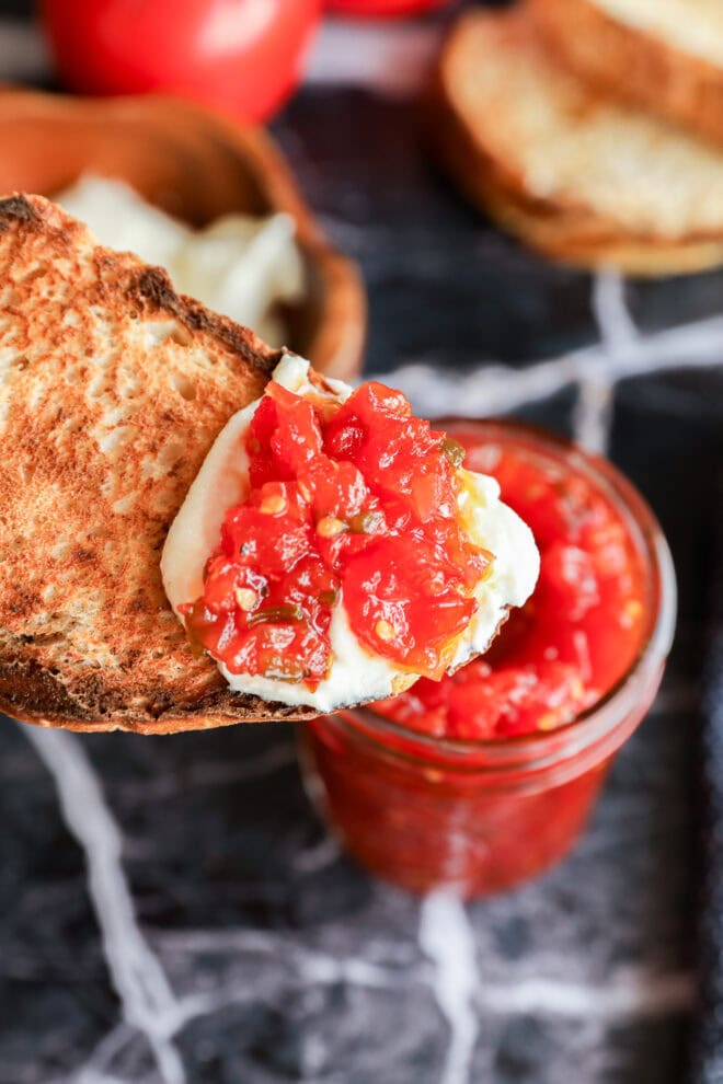 A piece of toasted bread topped with fresh mozzarella cheese and a spoonful of tomato relish is held above an open jar of tomato jam on a dark marble surface.