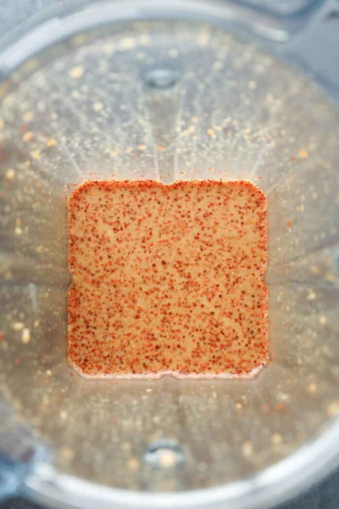 A close-up top view of a blender containing a tan liquid with small red specks, likely a blended sauce or dressing, with remnants clinging to the sides of the container.