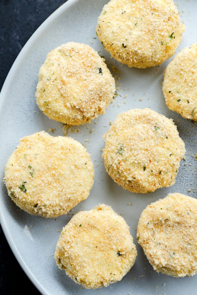 Seven breaded, round croquettes arranged on a light gray plate, viewed from above. The croquettes have a golden crust with small green herb flecks and a slightly rough, crumbly texture.