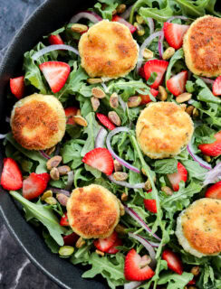 A salad with arugula, sliced strawberries, red onion, pistachios, and several breaded, golden-brown rounds of fried goat cheese, arranged in a black bowl on a dark marble surface.