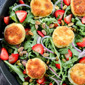 A salad with arugula, sliced strawberries, red onion, pistachios, and several breaded, golden-brown rounds of fried goat cheese, arranged in a black bowl on a dark marble surface.