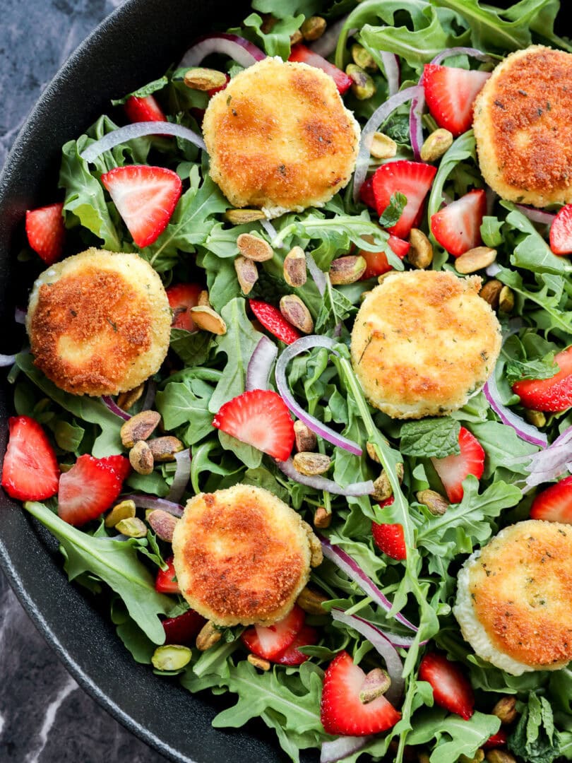 A salad with arugula, sliced strawberries, red onion, pistachios, and several breaded, golden-brown rounds of fried goat cheese, arranged in a black bowl on a dark marble surface.