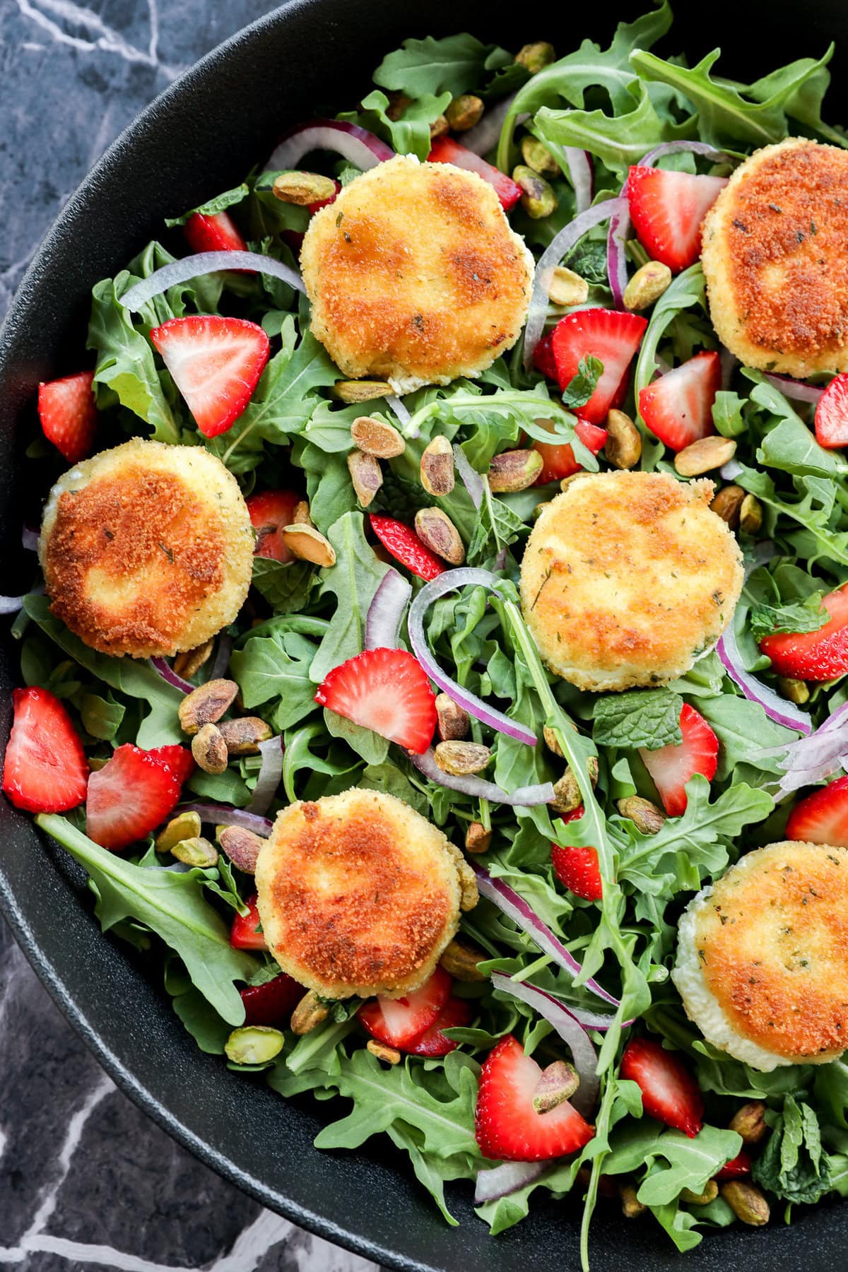 A salad with arugula, sliced strawberries, red onion, pistachios, and several breaded, golden-brown rounds of fried goat cheese, arranged in a black bowl on a dark marble surface.