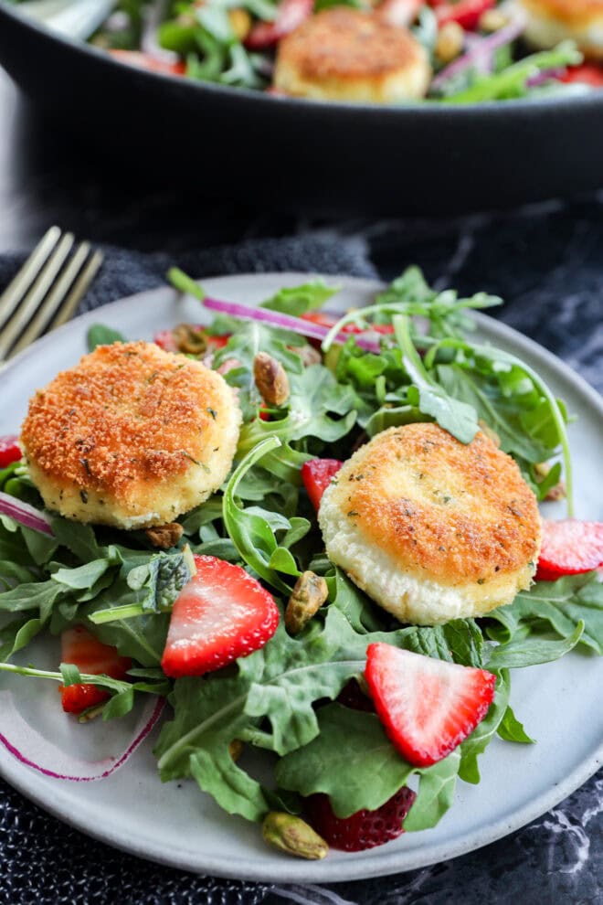 A plate of fresh arugula salad topped with sliced strawberries, roasted pistachios, and two golden-brown rounds of breaded goat cheese. A fork is beside the plate and more salad can be seen in the background.