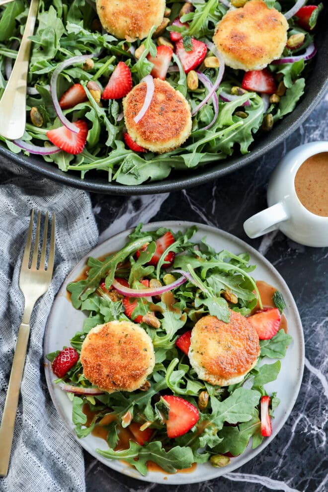 A fresh salad with arugula, sliced strawberries, red onions, pistachios, and crispy goat cheese rounds on a plate, with a fork, napkin, and small cup of dressing nearby.