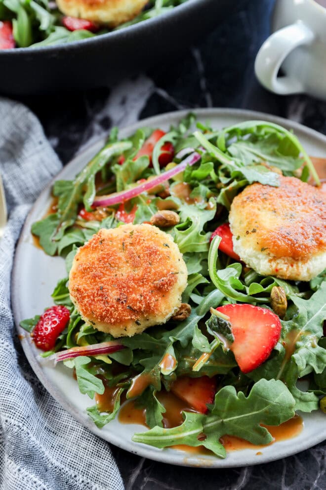 A plate of fresh arugula salad with sliced strawberries, red onions, pistachios, and two golden-brown breaded goat cheese rounds, drizzled with dressing.