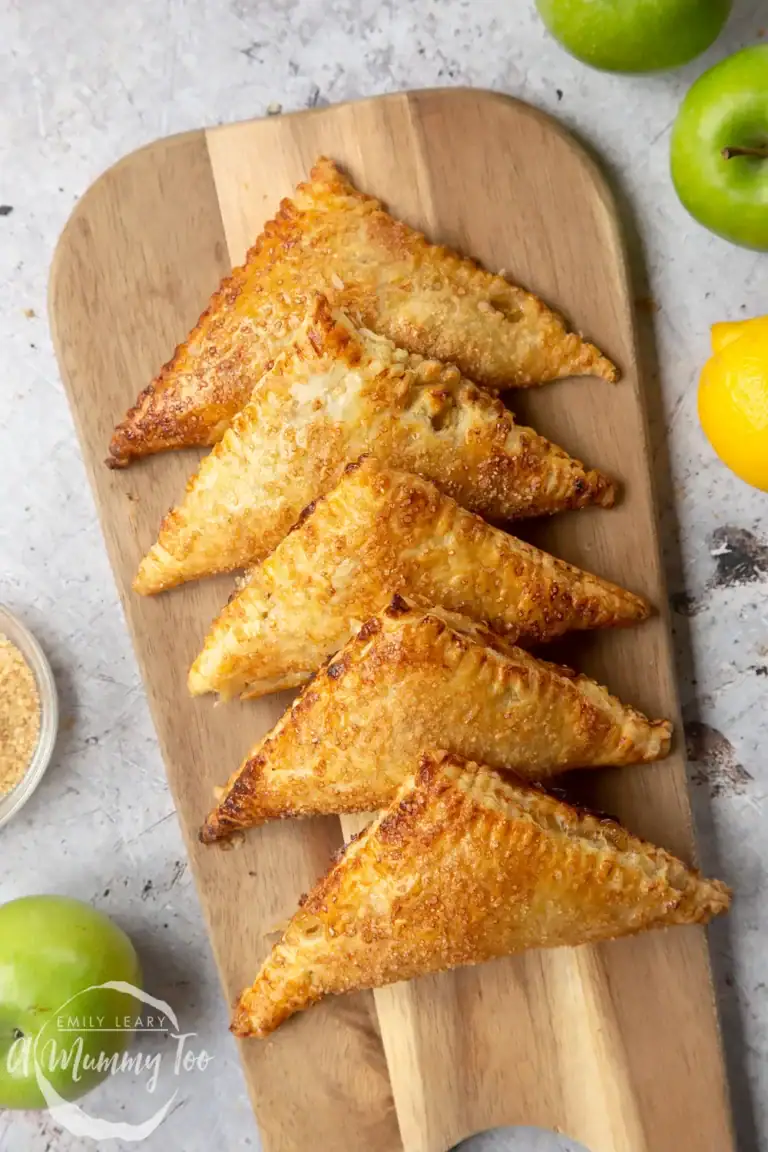 Five golden-brown puff pastry turnovers are arranged in a neat row on a wooden board, showcasing one of the tastiest puff pastry dessert recipes. Green apples, a lemon, and a small bowl of sugar sit around the board.