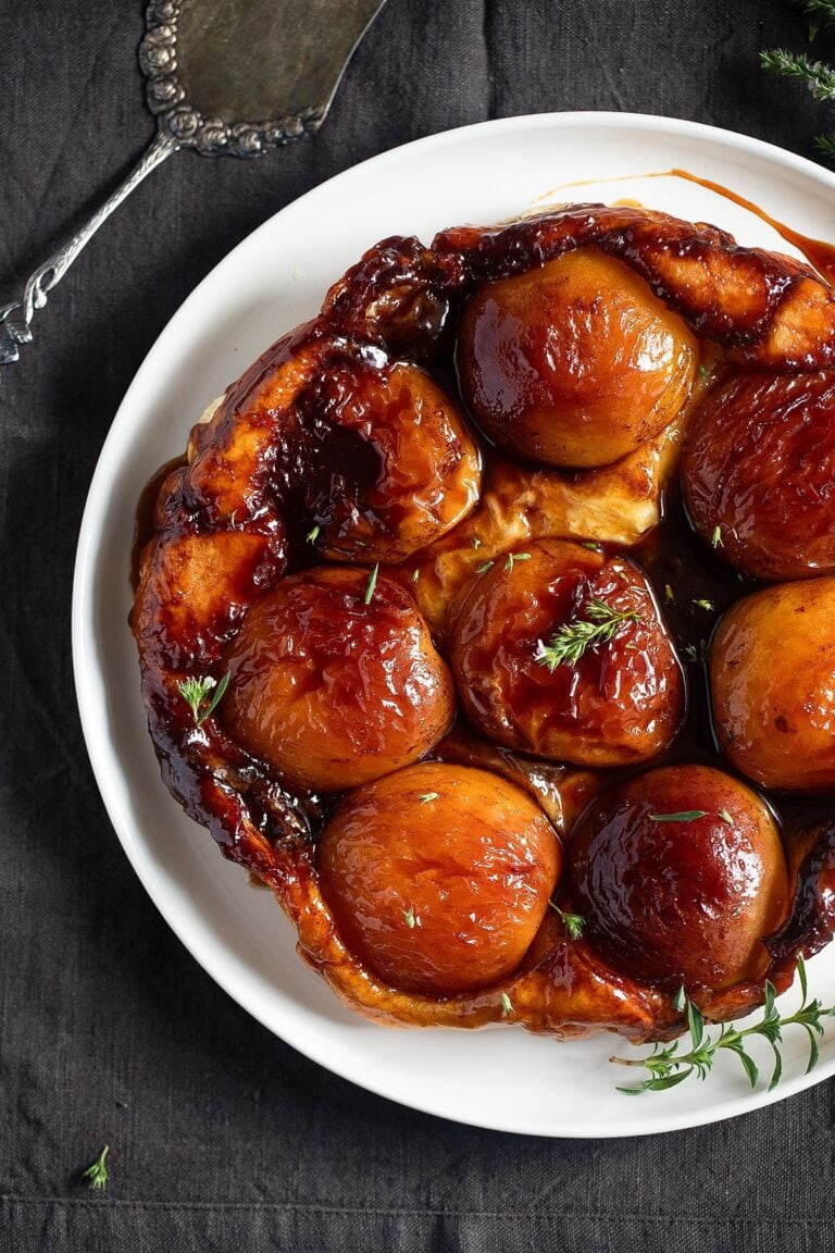 A close-up of an apple tarte Tatin with golden, caramelized apples on a flaky crust, garnished with fresh thyme—a standout among puff pastry dessert recipes—served on a white plate with a pie server on a dark tablecloth.