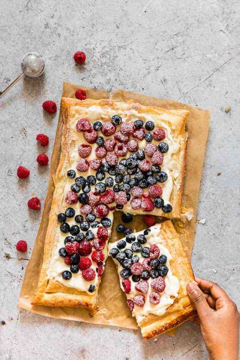 A hand holds a slice of a rectangular fruit tart topped with raspberries and blueberries on cream cheese over golden puff pastry, an inviting idea for those seeking new puff pastry dessert recipes, with loose berries scattered nearby.