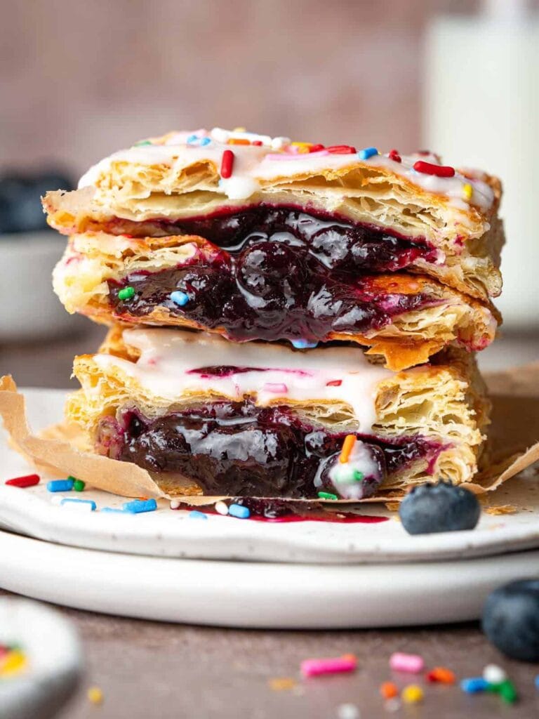 A close-up of two stacked blueberry pastries with flaky crusts, gooey blueberry filling, white icing, and colorful sprinkles—an irresistible treat inspired by puff pastry dessert recipes—served on a white plate with blueberries and extra sprinkles.