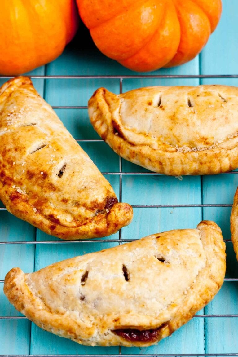 Four baked pumpkin hand pies with golden crusts rest on a cooling rack, showcasing one of the best puff pastry dessert recipes, with two small orange pumpkins in the background on a bright turquoise surface.
