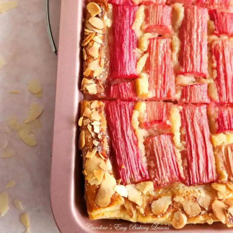 A close-up of a baked tart in a pan, topped with rows of pink rhubarb stalks and sliced almonds on the crust. Reminiscent of classic puff pastry dessert recipes, the tart is cut into neat squares with almond slices scattered nearby.