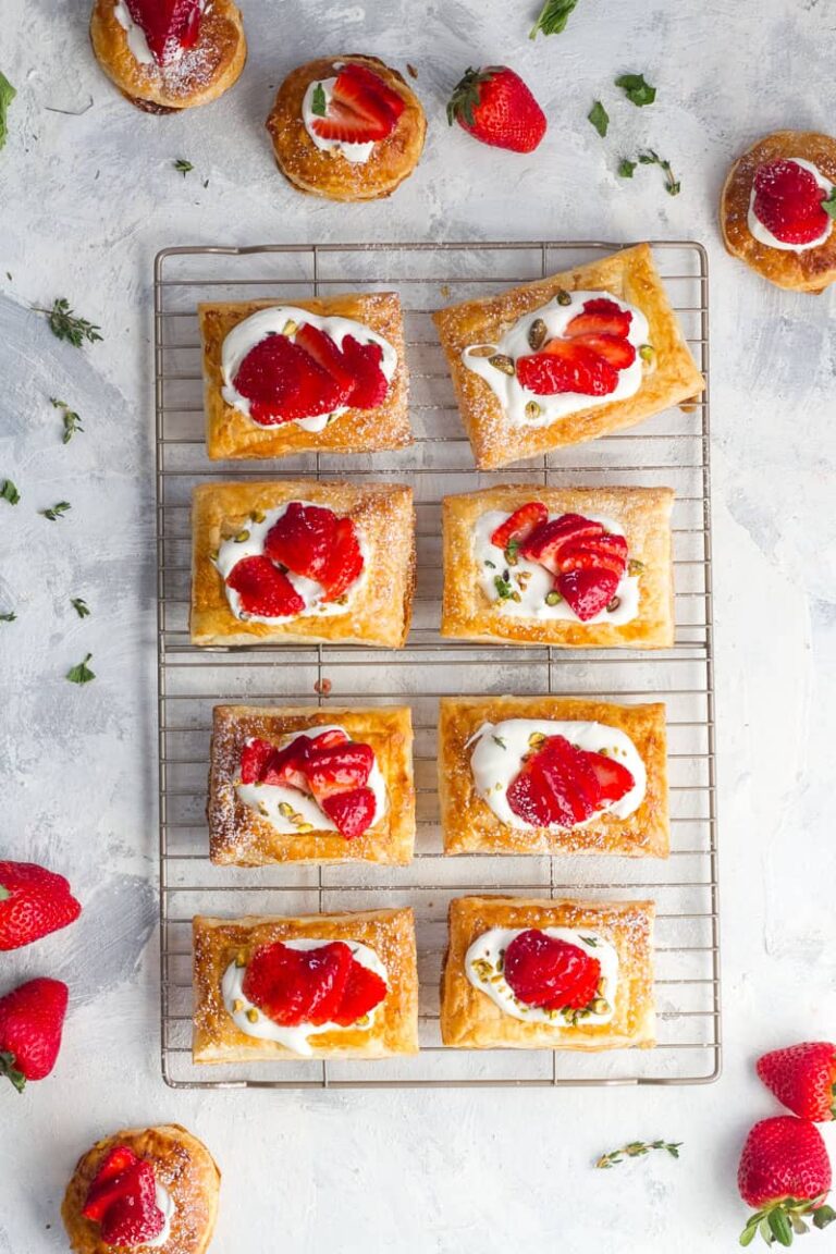 Rectangular puff pastry tarts topped with whipped cream and sliced strawberries are arranged on a cooling rack, surrounded by extra strawberries—an enticing addition to your collection of puff pastry dessert recipes.