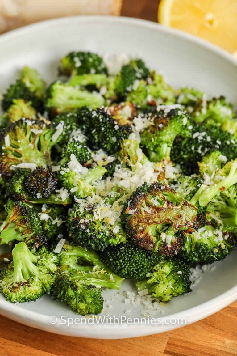 A white bowl filled with roasted broccoli florets topped with grated Parmesan cheese, with part of a lemon visible in the background.
