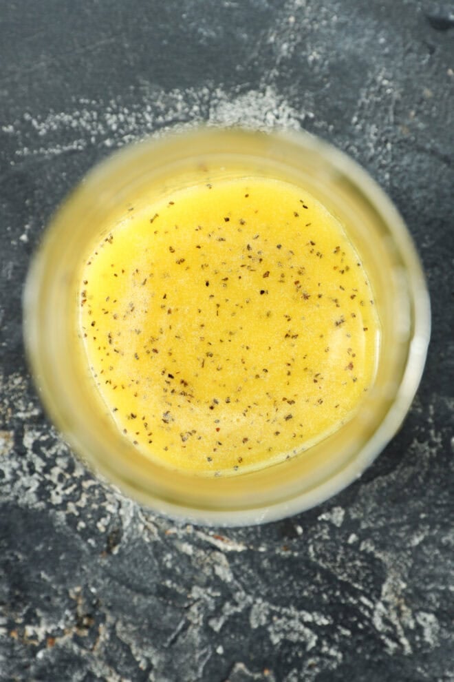 A close-up, top-down view of a glass jar filled with yellow salad dressing speckled with black pepper, set on a dark textured surface—perfect for drizzling.