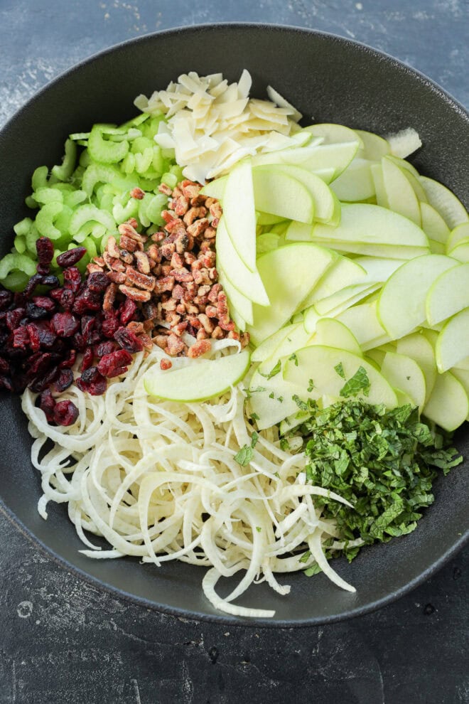 A bowl filled with sliced green apples, chopped celery, slivered almonds, dried cranberries, chopped pecans, spiralized fennel, and fresh herbs creates a refreshing Apple Fennel Salad bursting with flavor and crunch.